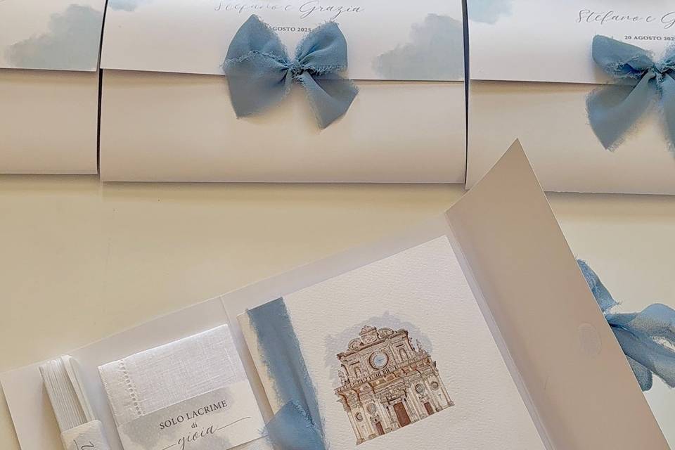Partecipazioni arrotolate e chiuse con un fiocco di seta azzurra sfrangiata, e sotto una cartella aperta con dentro un acquerello dipinto a mano della facciata di una chiesa barocca