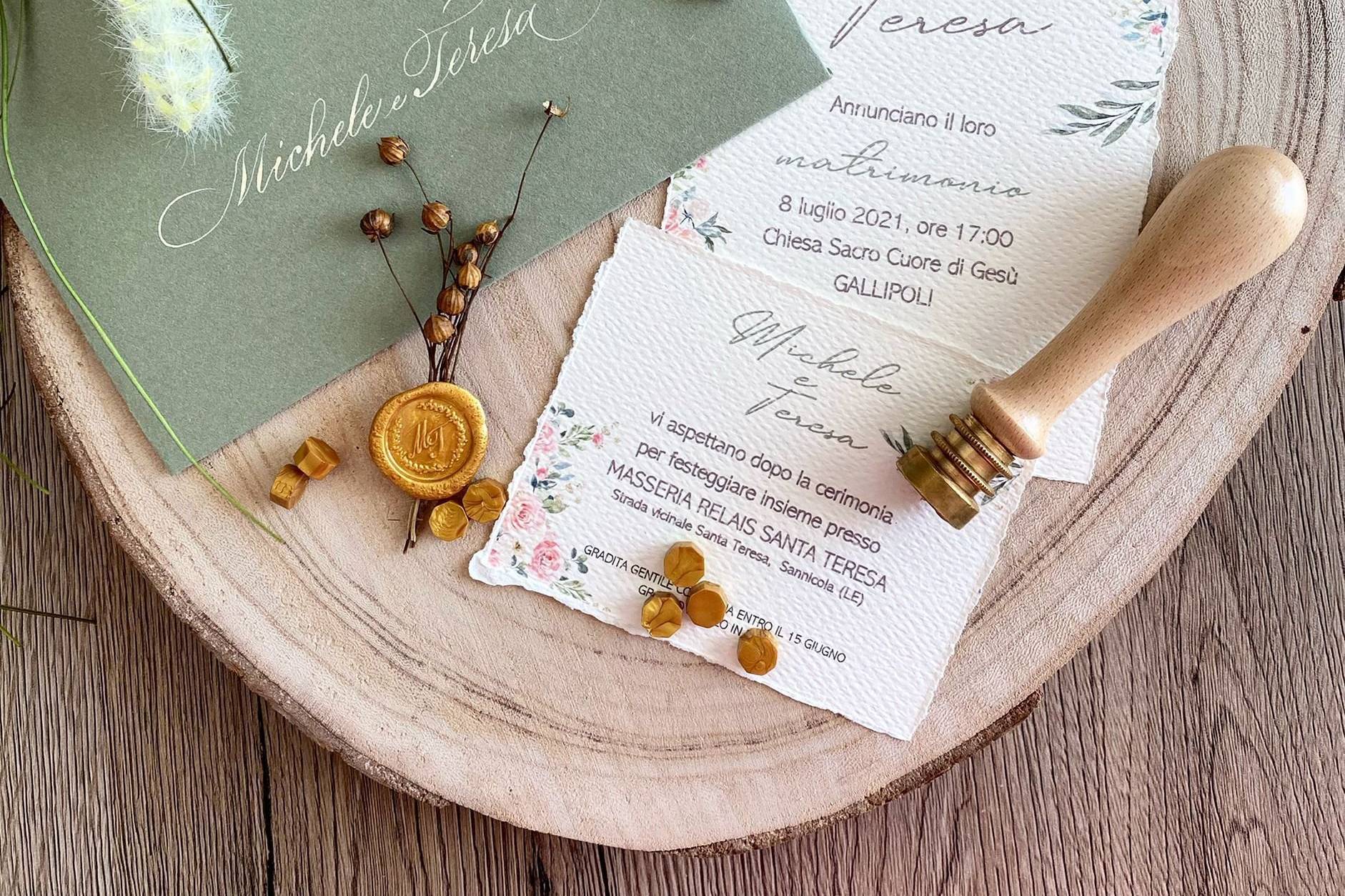 Partecipazioni di nozze su carta a mano dal bordo sfrangiato, stampate con acquerelli floreali, accanto a una busta verde salvia, a un sigillo di ceralacca dorata e al timbro di legno usato per imprimerlo