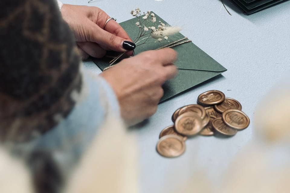 Sara al banco, ripresa da dietro le spalle: le mani legano con lo spago un mazzetto di fiori secchi sopra una busta verde salvia, e accanto sul piano c'è una pila di sigilli di ceralacca dorata già pronti